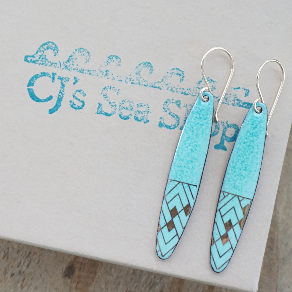 a pair of seafoam green enamel earrings with gold designs, displayed on a gift box