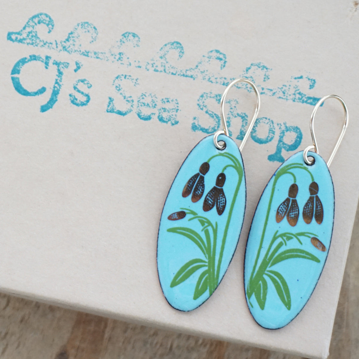 a pair of light blue enamel oval shaped earrings with gold flower accents, displayed on a gift box