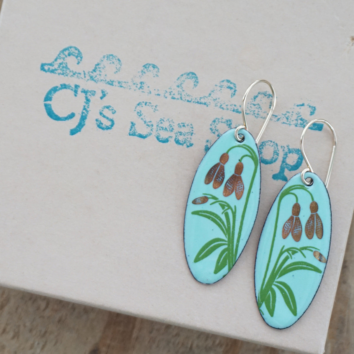a pair of seafoam green enamel oval shaped earrings with gold flower accents, displayed on a gift box