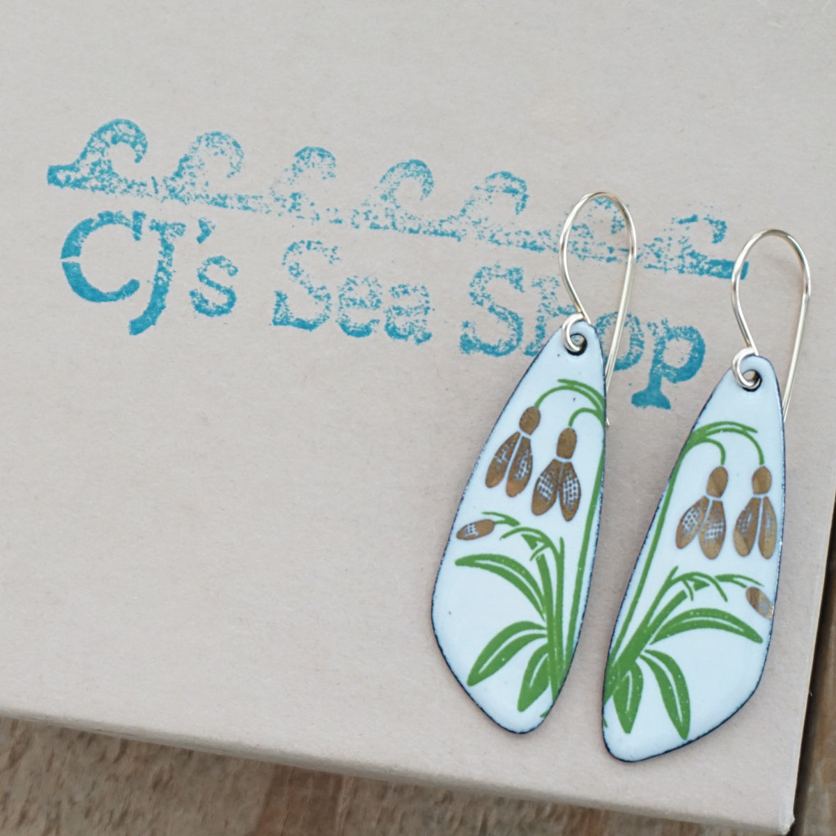 a pair of white enamel teardrop shaped earrings with gold flower accents, displayed on a gift box