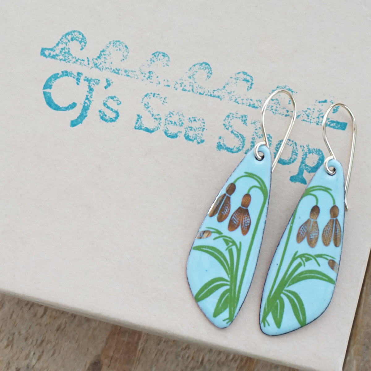a pair of light blue enamel teardrop shaped earrings with gold flower accents, displayed on a gift box