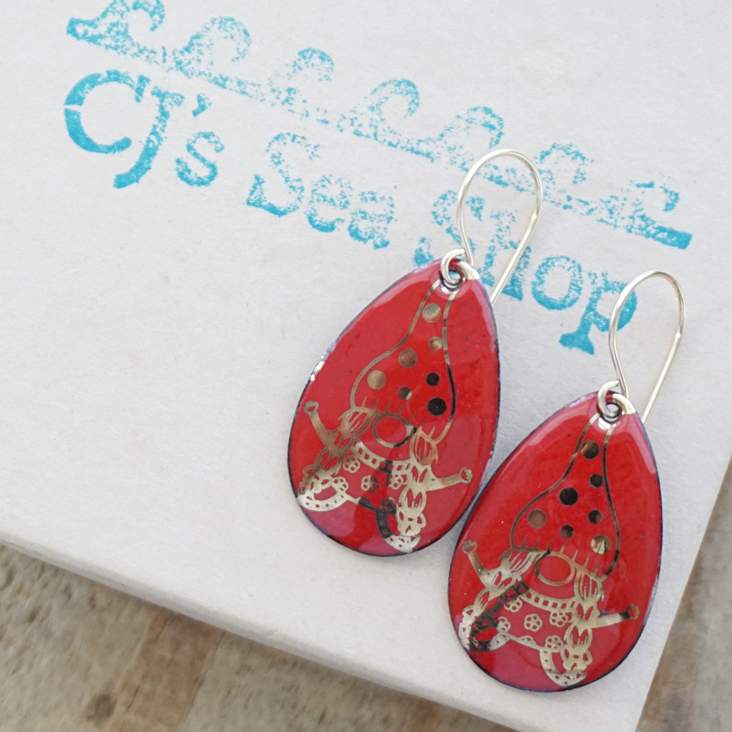 A pair of red enamel teardrop earrings with gold gnome designs, on a gift box