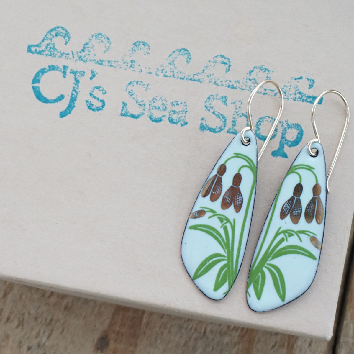 a pair of light green enamel teardrop shaped earrings with gold flower accents, displayed on a gift box