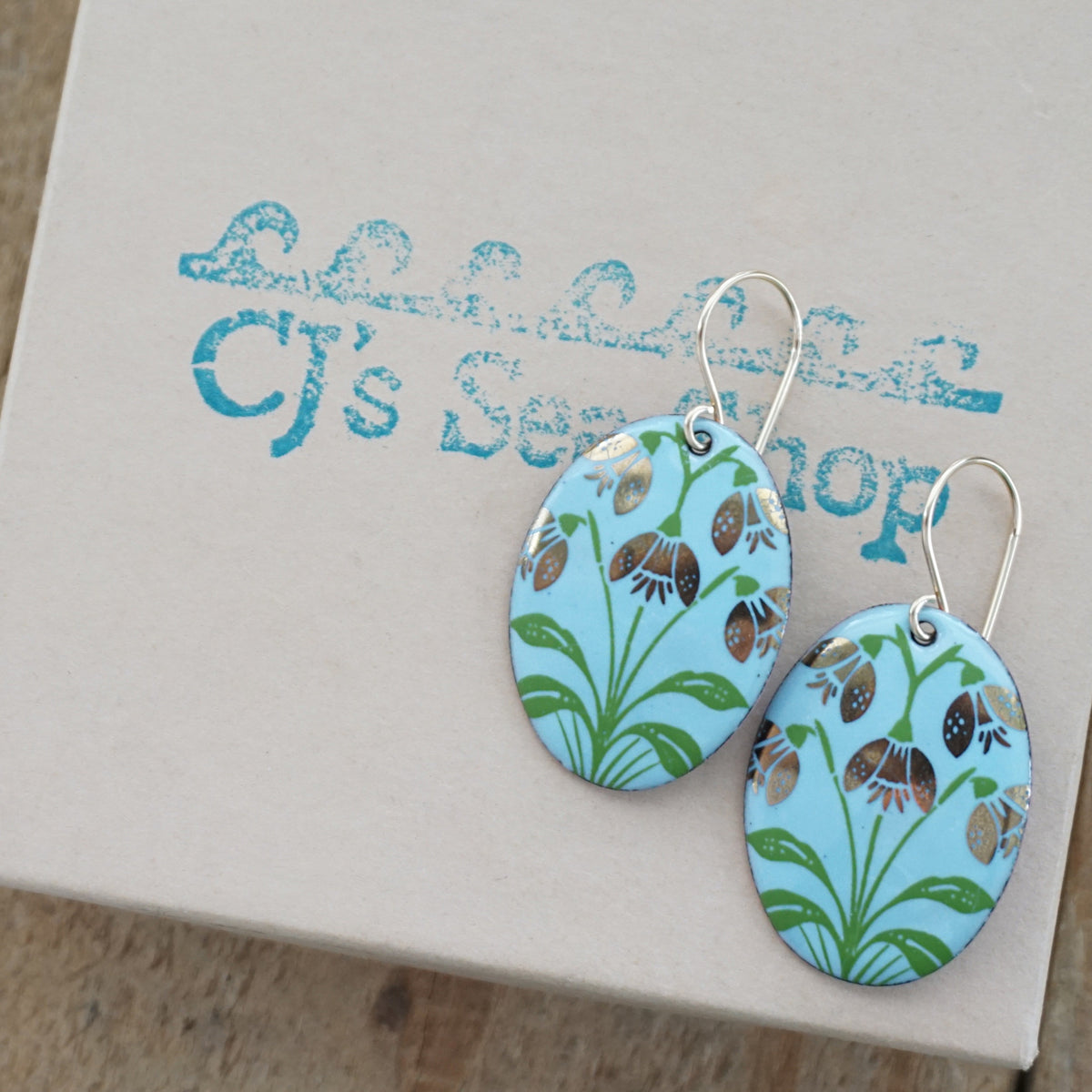a pair of light blue enamel oval shaped earrings with gold flower accents, displayed on a gift box