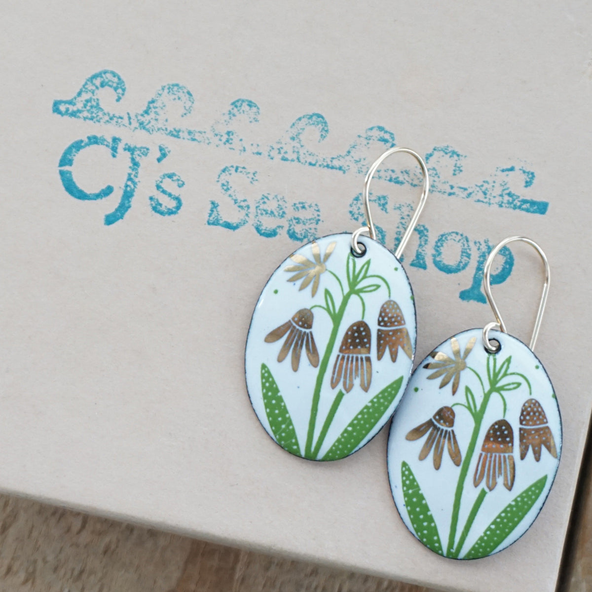 a pair of white enamel oval shaped earrings with gold flower accents, displayed on a gift box
