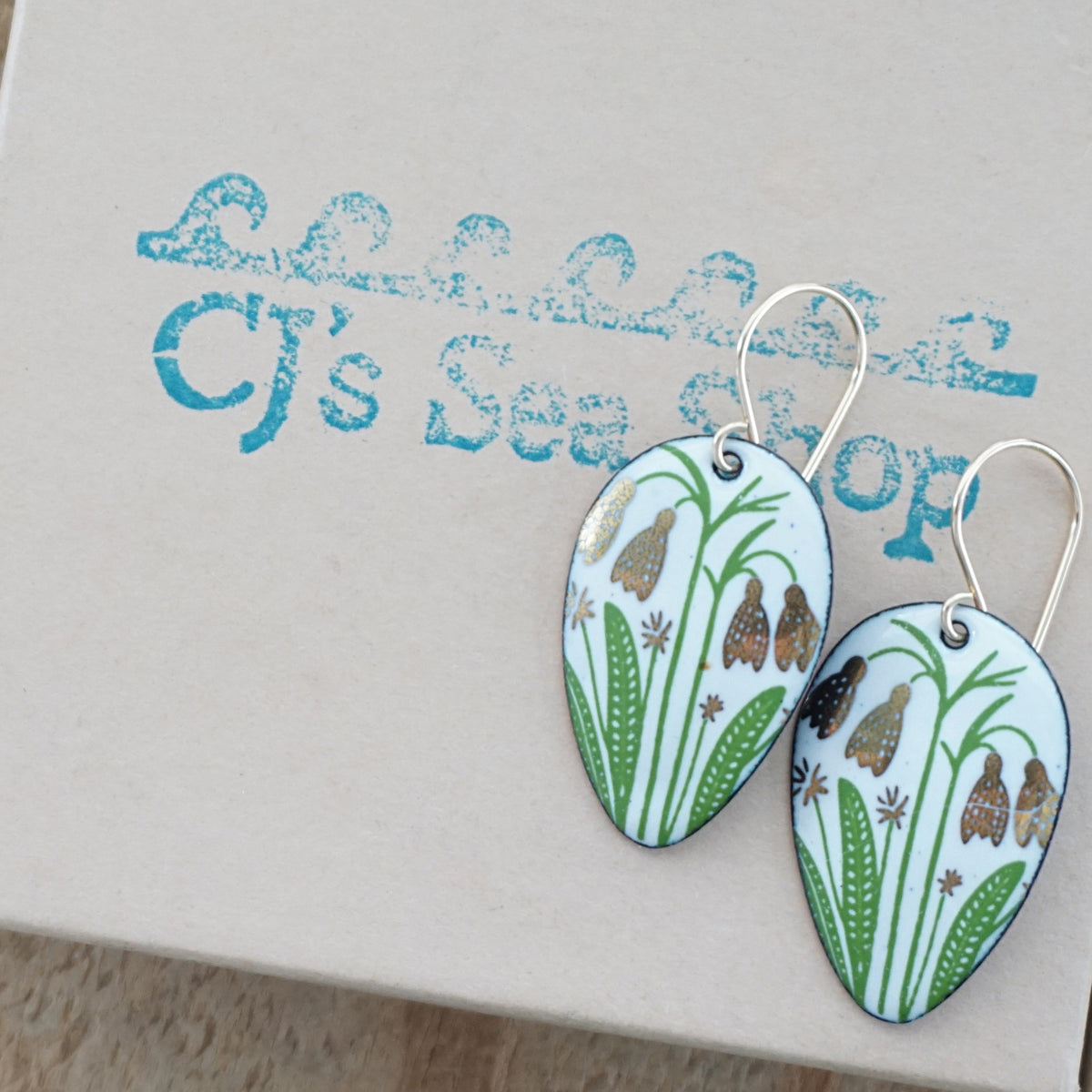 a pair of white enamel teardrop shaped earrings with gold flower accents, displayed on a gift box