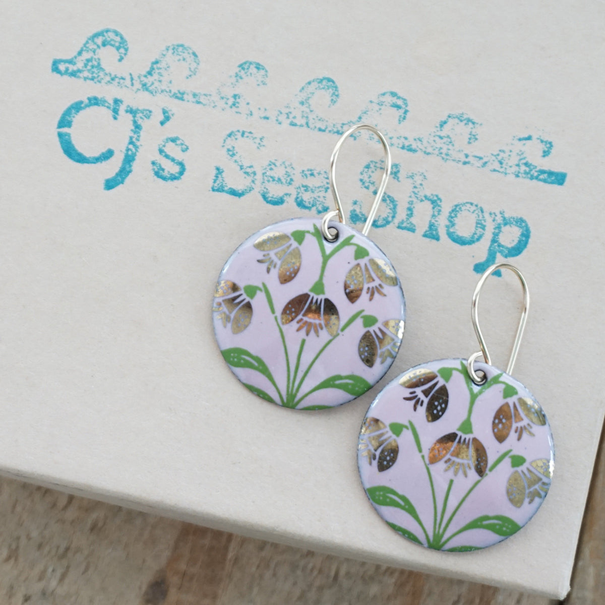 a pair of pink enamel disc shaped earrings with gold flower accents, displayed on a gift box