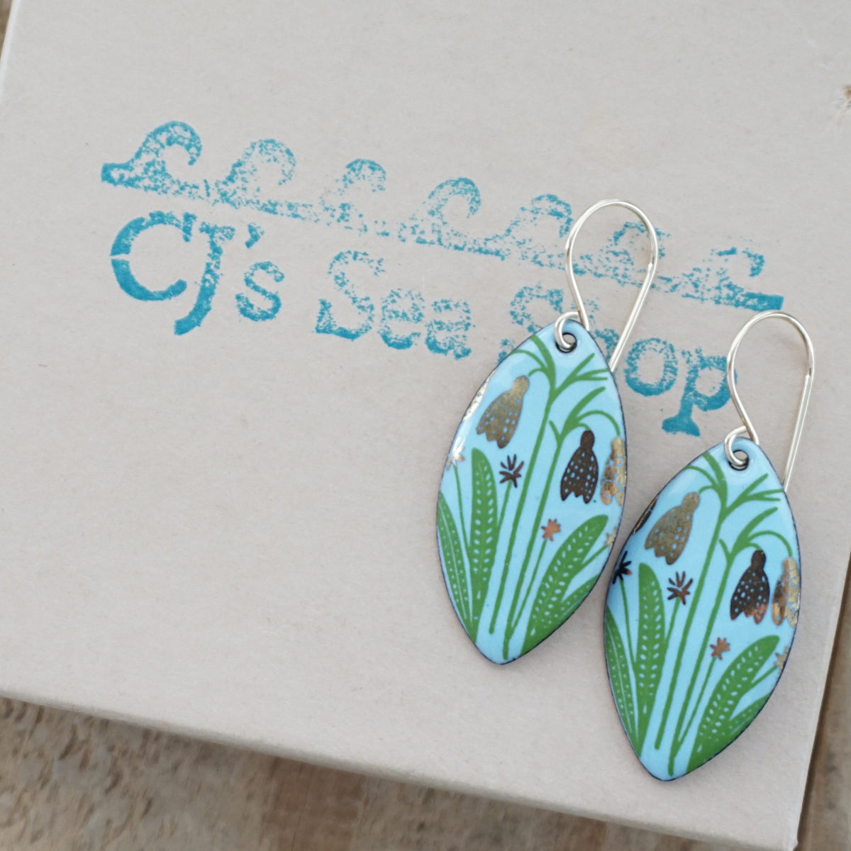 a pair of light blue enamel marquis shaped earrings with gold flower accents, displayed on a gift box
