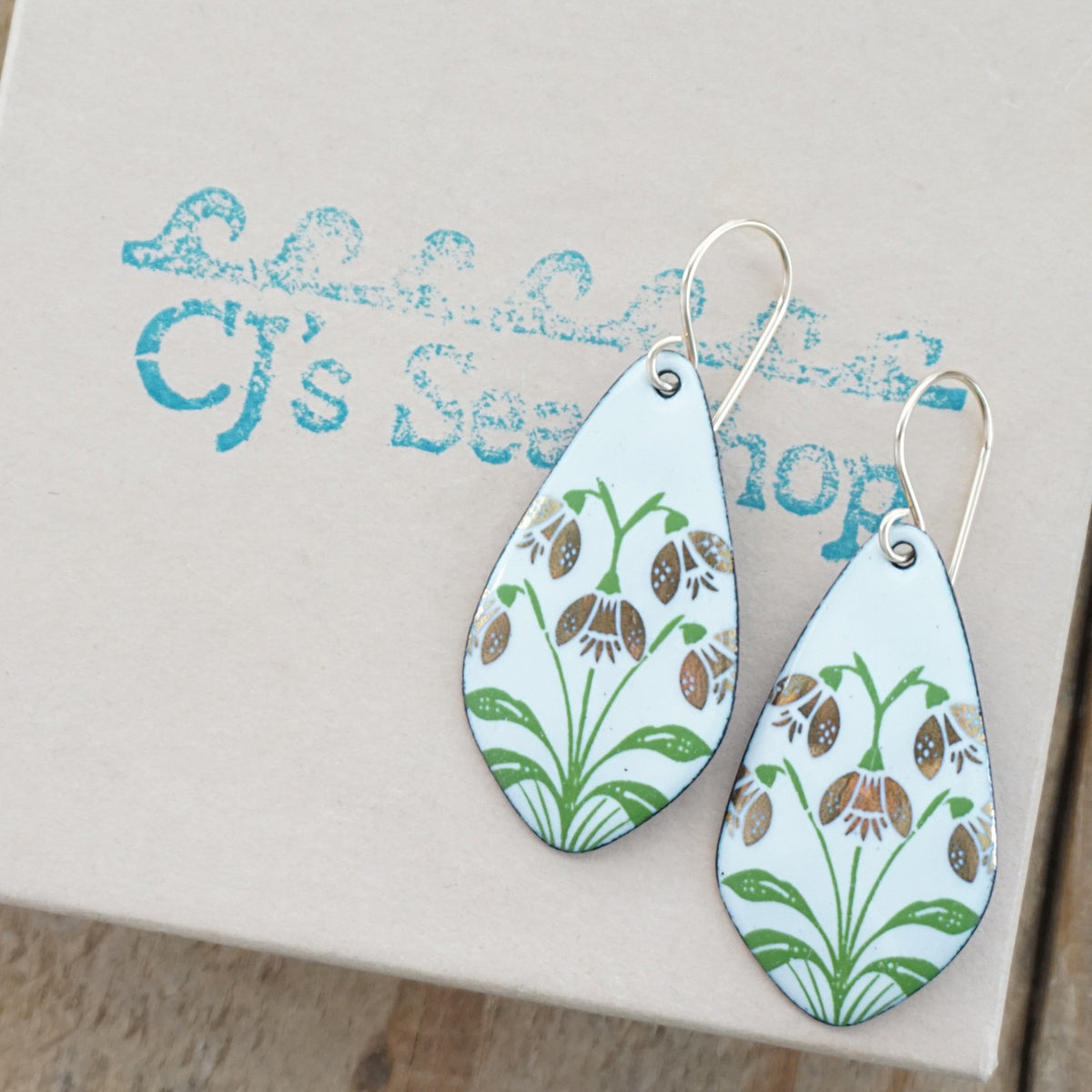 a pair of white enamel teardrop shaped earrings with gold flower accents, displayed on a gift box