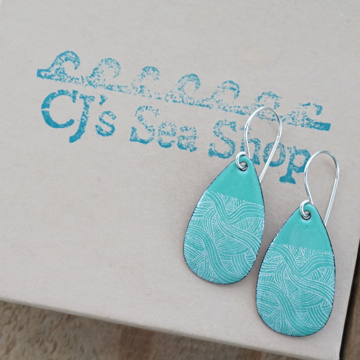 a pair of green enamel teardrop shaped earrings with white wavy accents, displayed on a gift box