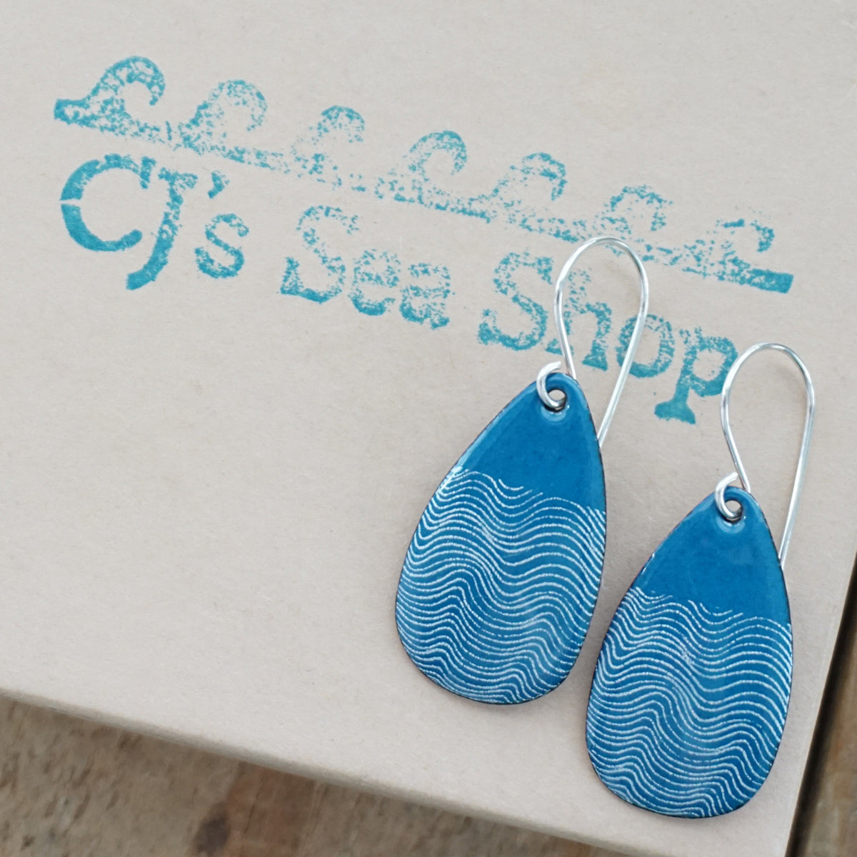 a pair of teal blue enamel teardrop shaped earrings with white wavy accents, displayed on a gift box