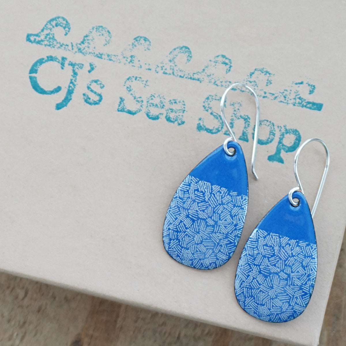 a pair of blue enamel teardrop shaped earrings with white accents, displayed on a gift box