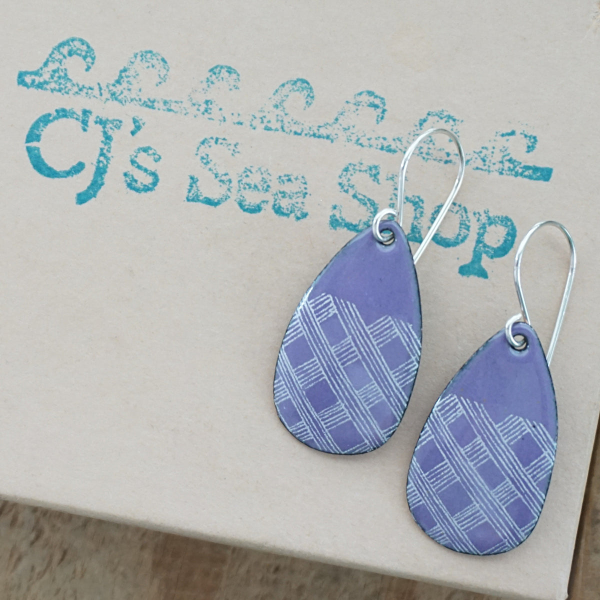 a pair of purple enamel teardrop shaped earrings with white gingham accents, displayed on a gift box