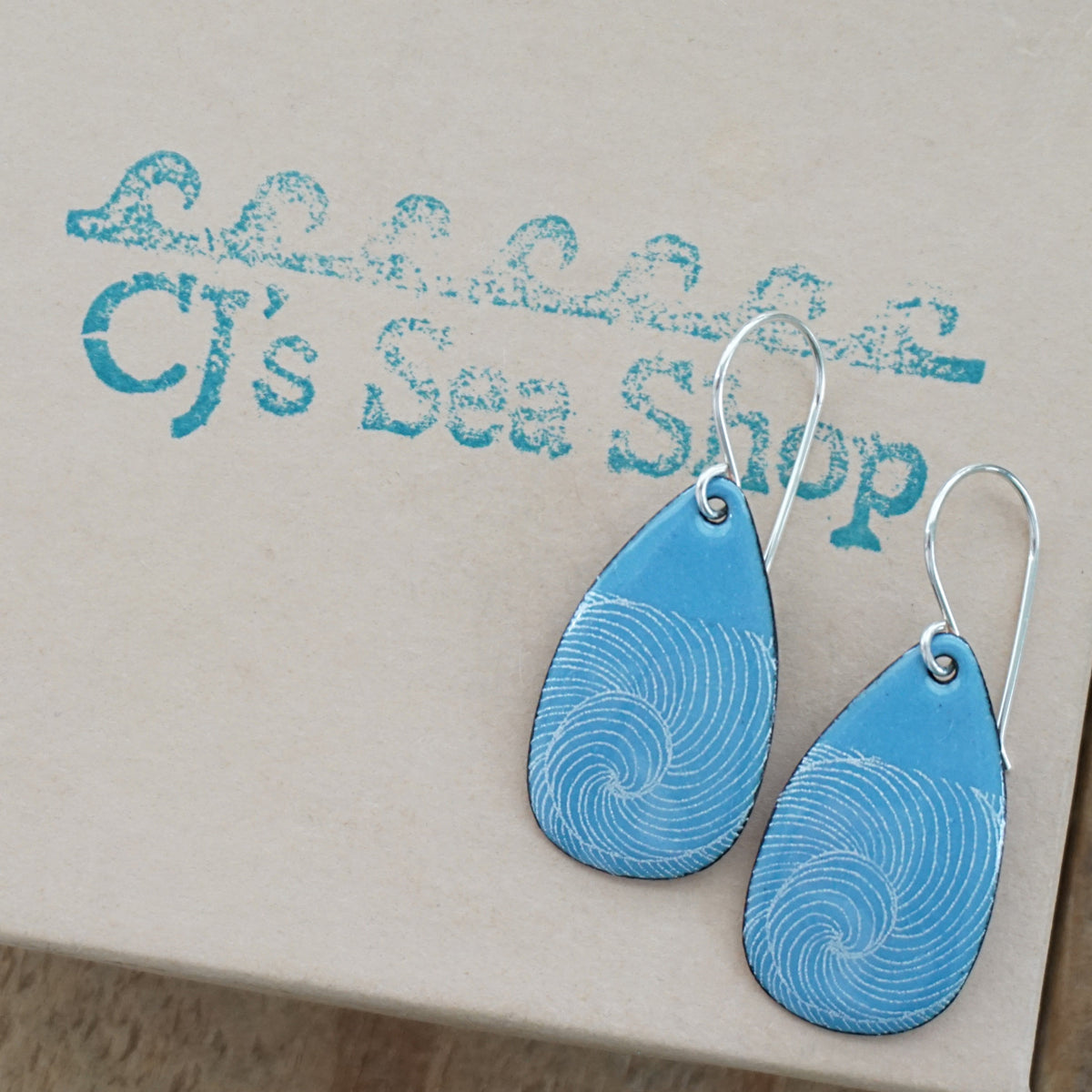 a pair of blue enamel teardrop shaped earrings with white wave accents, displayed on a gift box