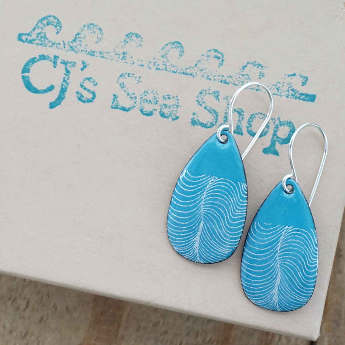 a pair of blue enamel teardrop shaped earrings with white wavy accents, displayed on a gift box