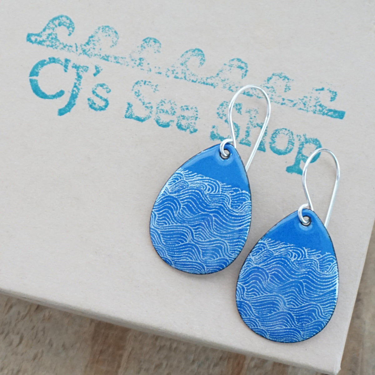 a pair of blue enamel teardrop shaped earrings with white wave accents, displayed on a gift box