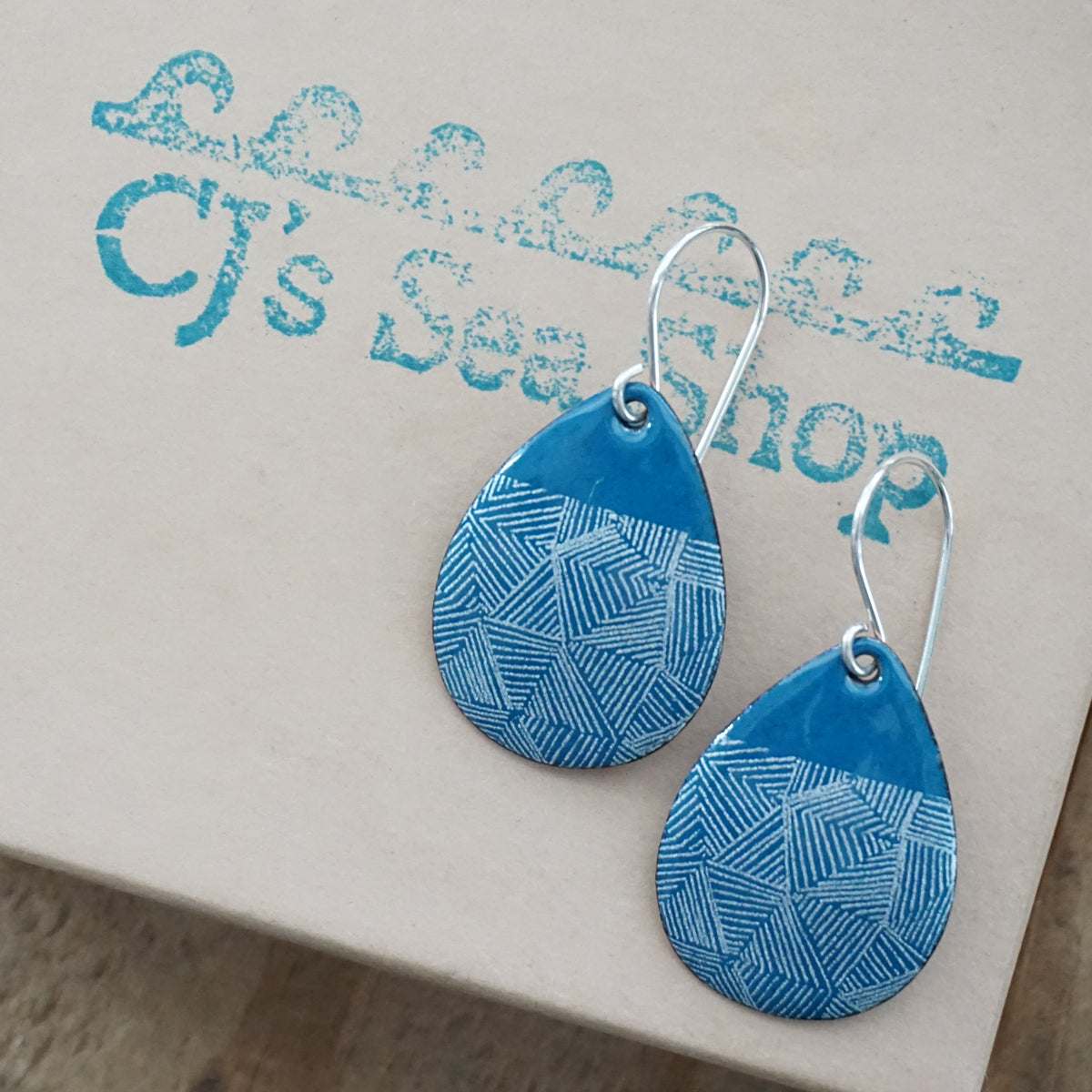 a pair of teal blue enamel teardrop shaped earrings with white geometric accents, displayed on a gift box