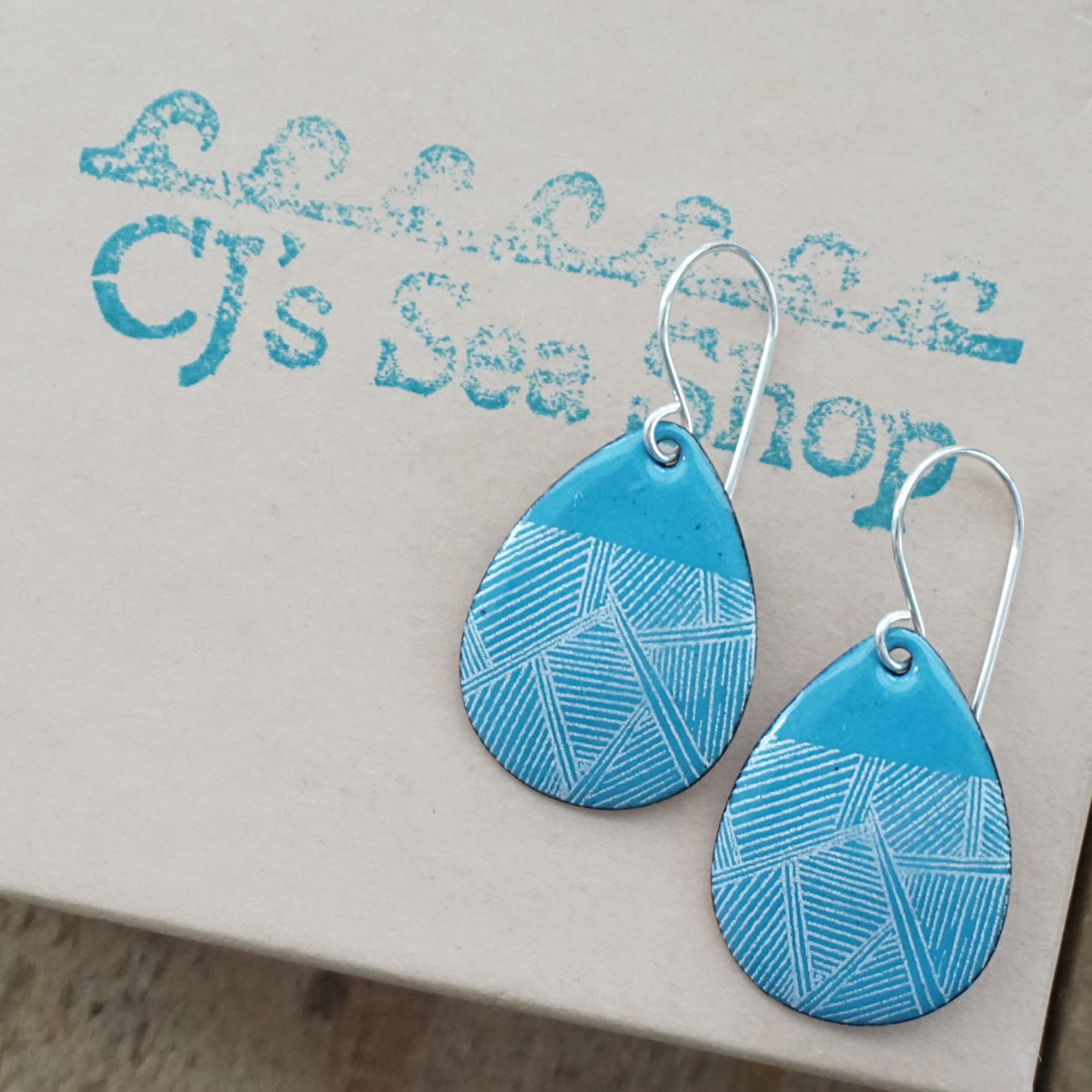 a pair of blue enamel teardrop shaped earrings with white geometric accents, displayed on a gift box