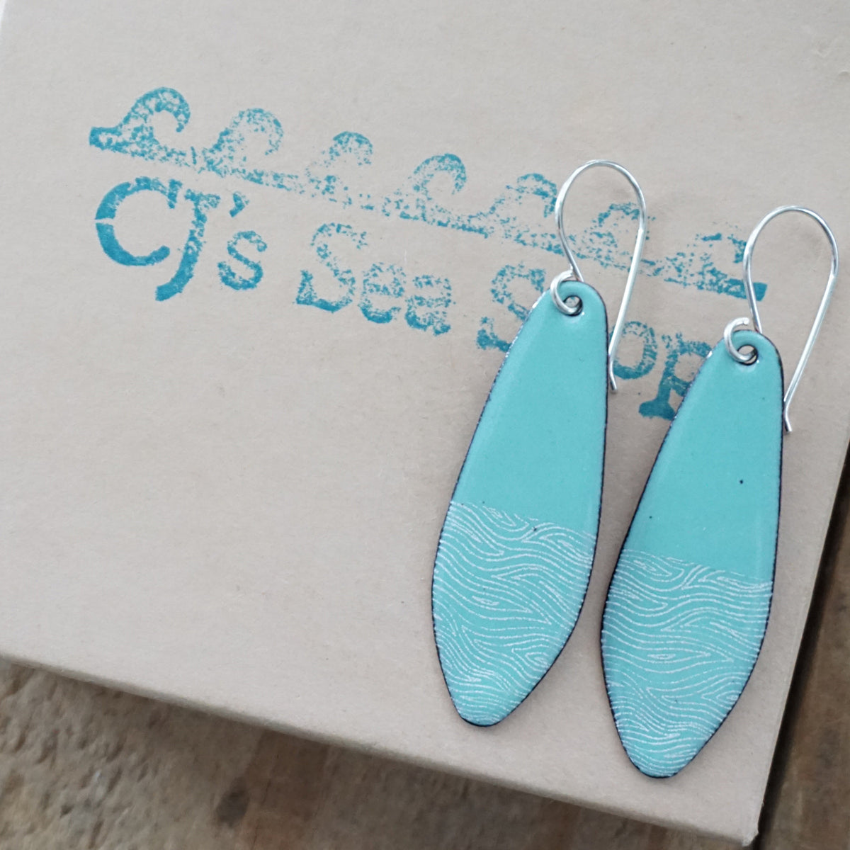 a pair of light green enamel teardrop shaped earrings with white wavy accents, displayed on a gift box