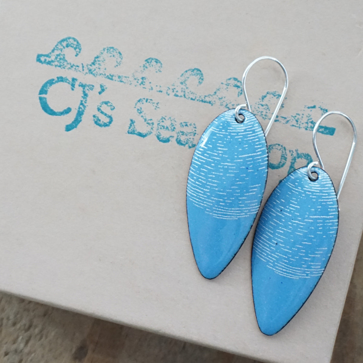 a pair of blue enamel teardrop shaped earrings with white accents, displayed on a gift box