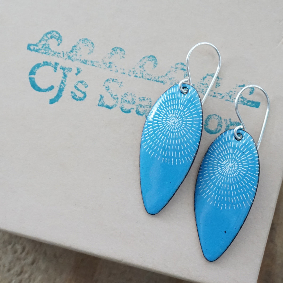a pair of blue enamel teardrop shaped earrings with white accents, displayed on a gift box