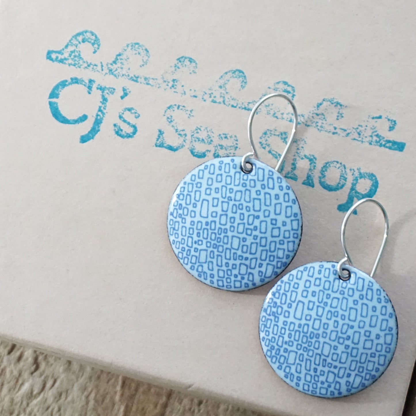 A pair of light seafoam green enamel disc earrings with blue square accents on a gift box