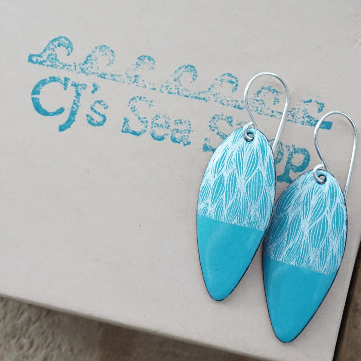 a pair of teal green enamel teardrop shaped earrings with white leaf accents, displayed on a gift box