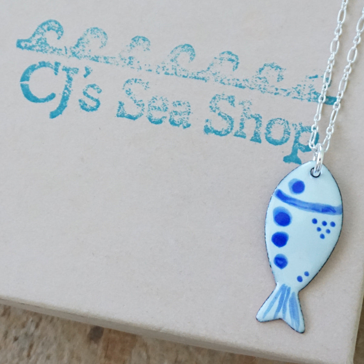 light green and blue enamel fish shaped pendant, displayed on a gift box