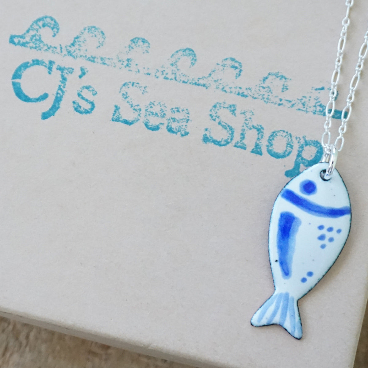 white and blue enamel fish shaped pendant, displayed on a gift box