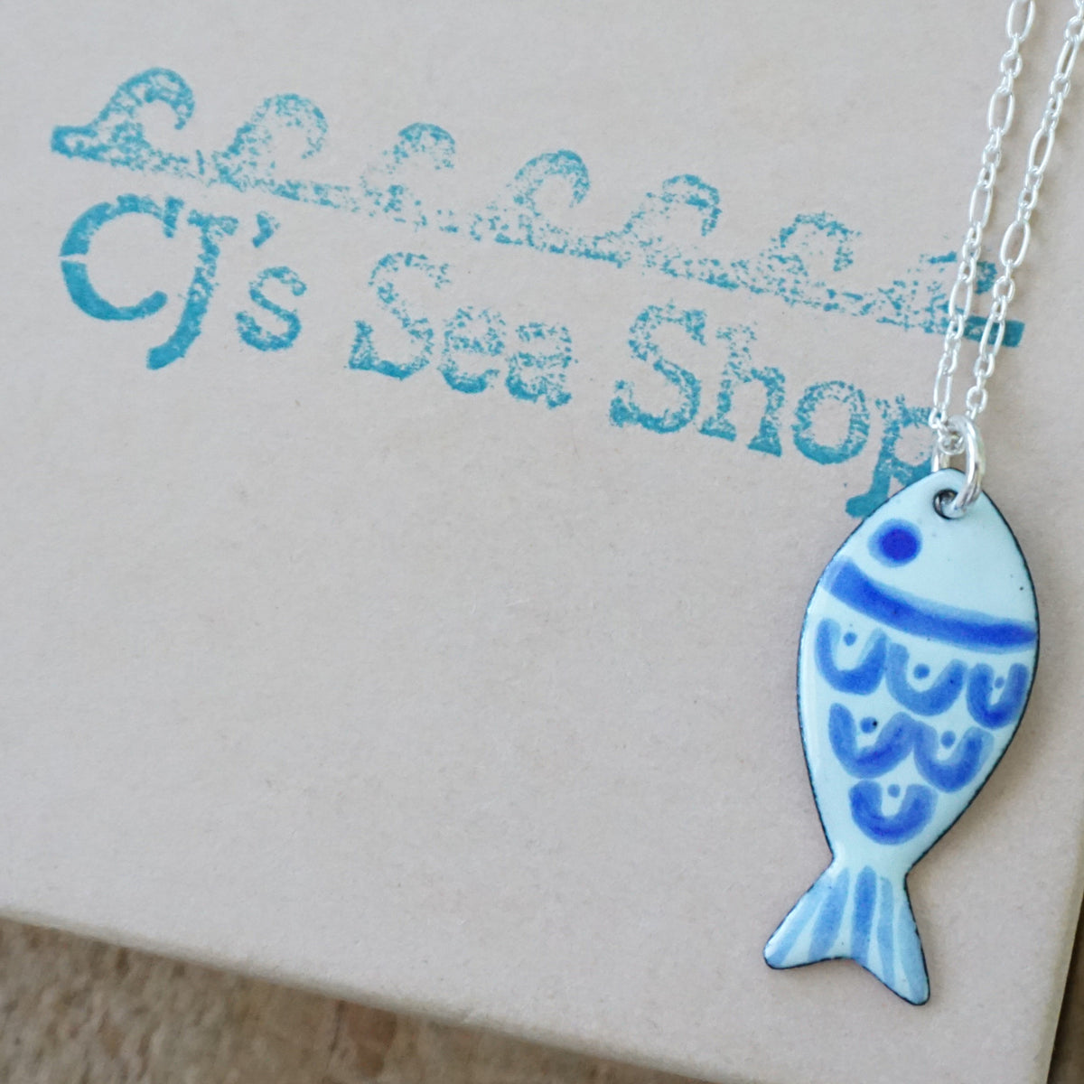 light green and blue enamel fish shaped pendant, displayed on a gift box