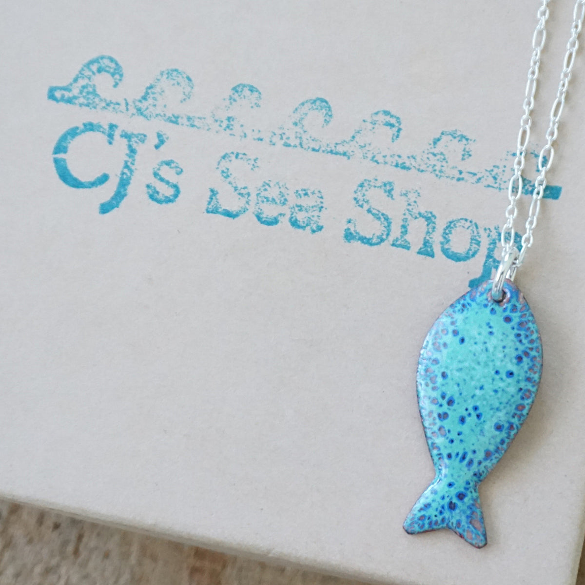 green and blue enamel fish shaped pendant, displayed on a gift box