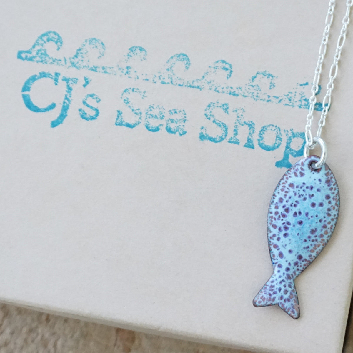 light blue and purple enamel fish shaped pendant against a wooden background