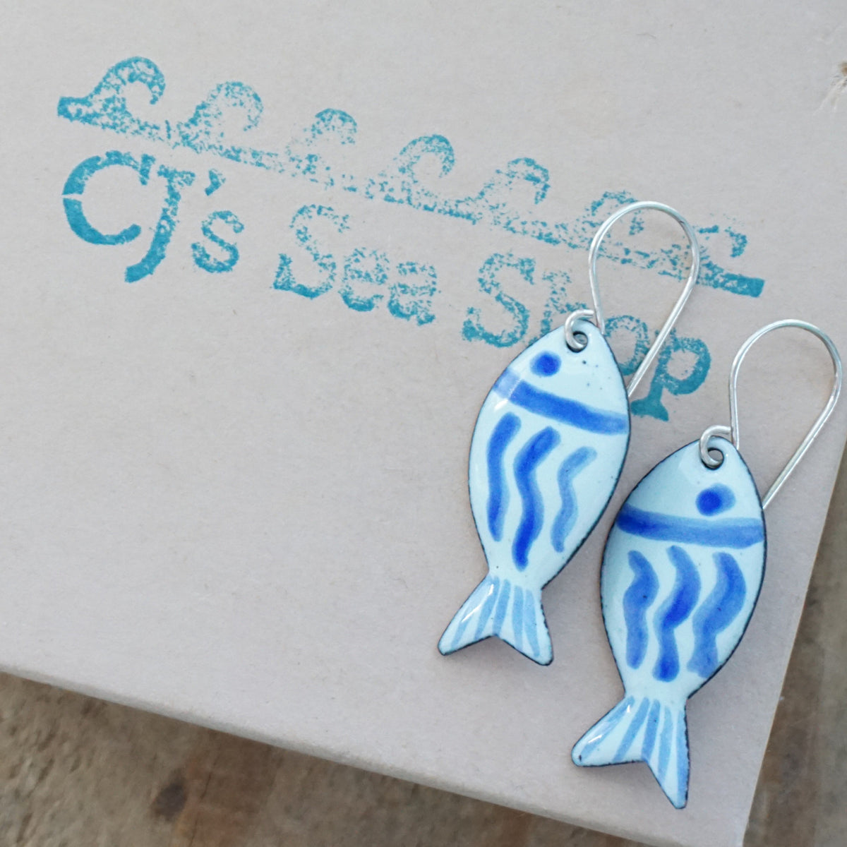 light green and blue enamel fish shaped earrings, displayed on a gift box