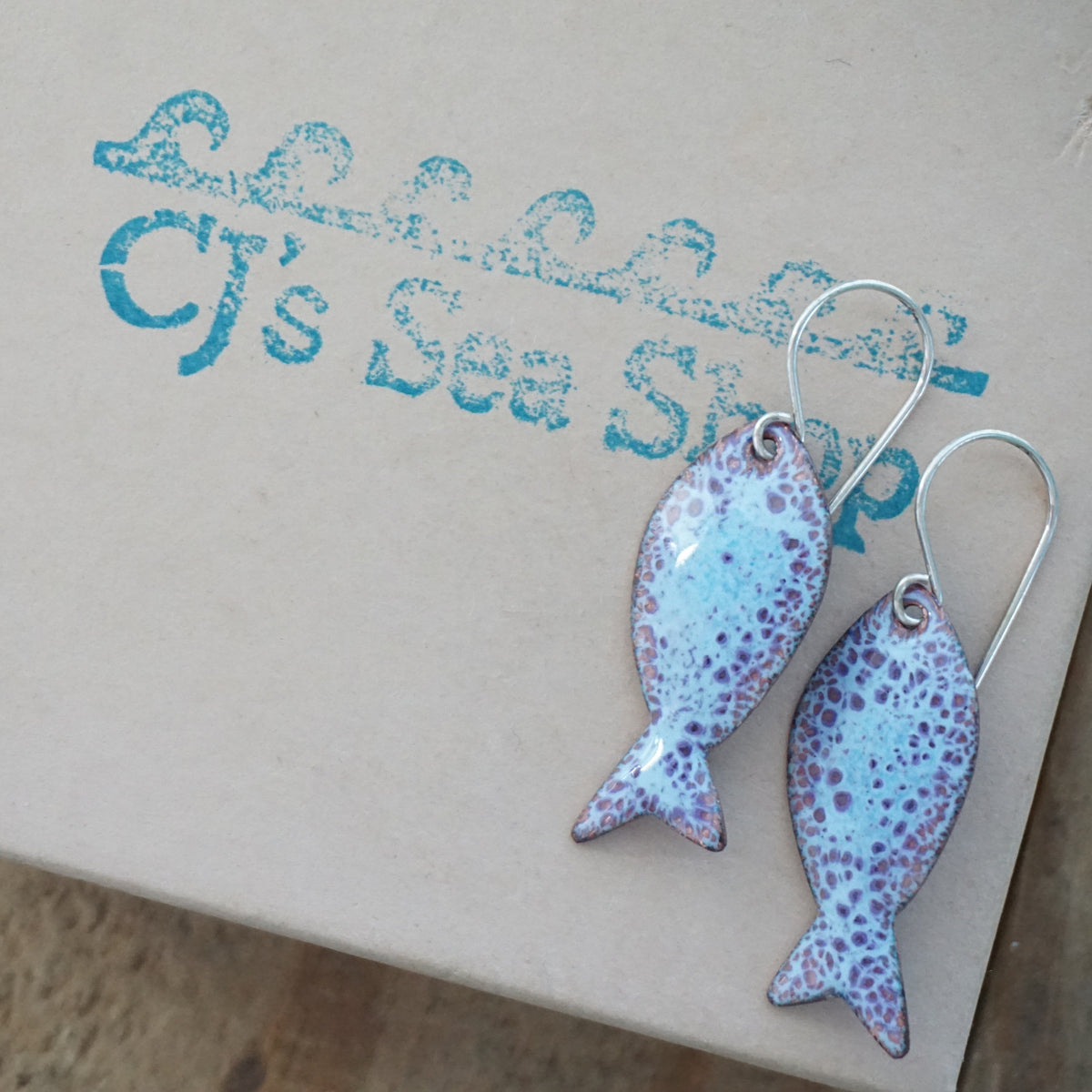 light blue and purple enamel fish shaped earrings, displayed on a gift box
