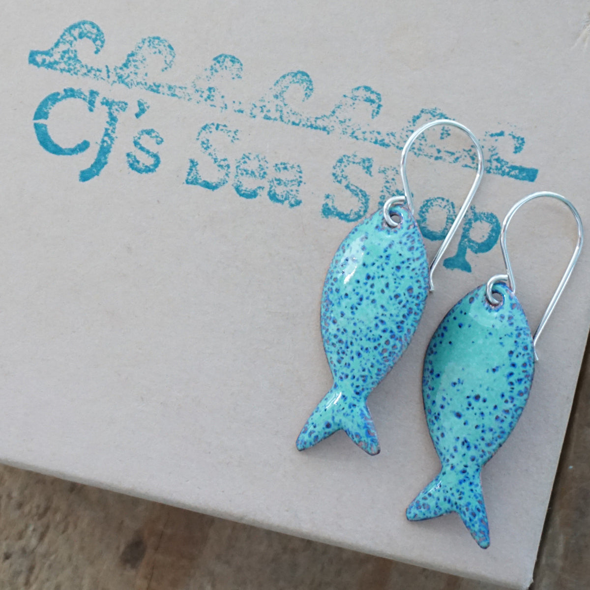 green and blue enamel fish shaped earrings, displayed on a gift box