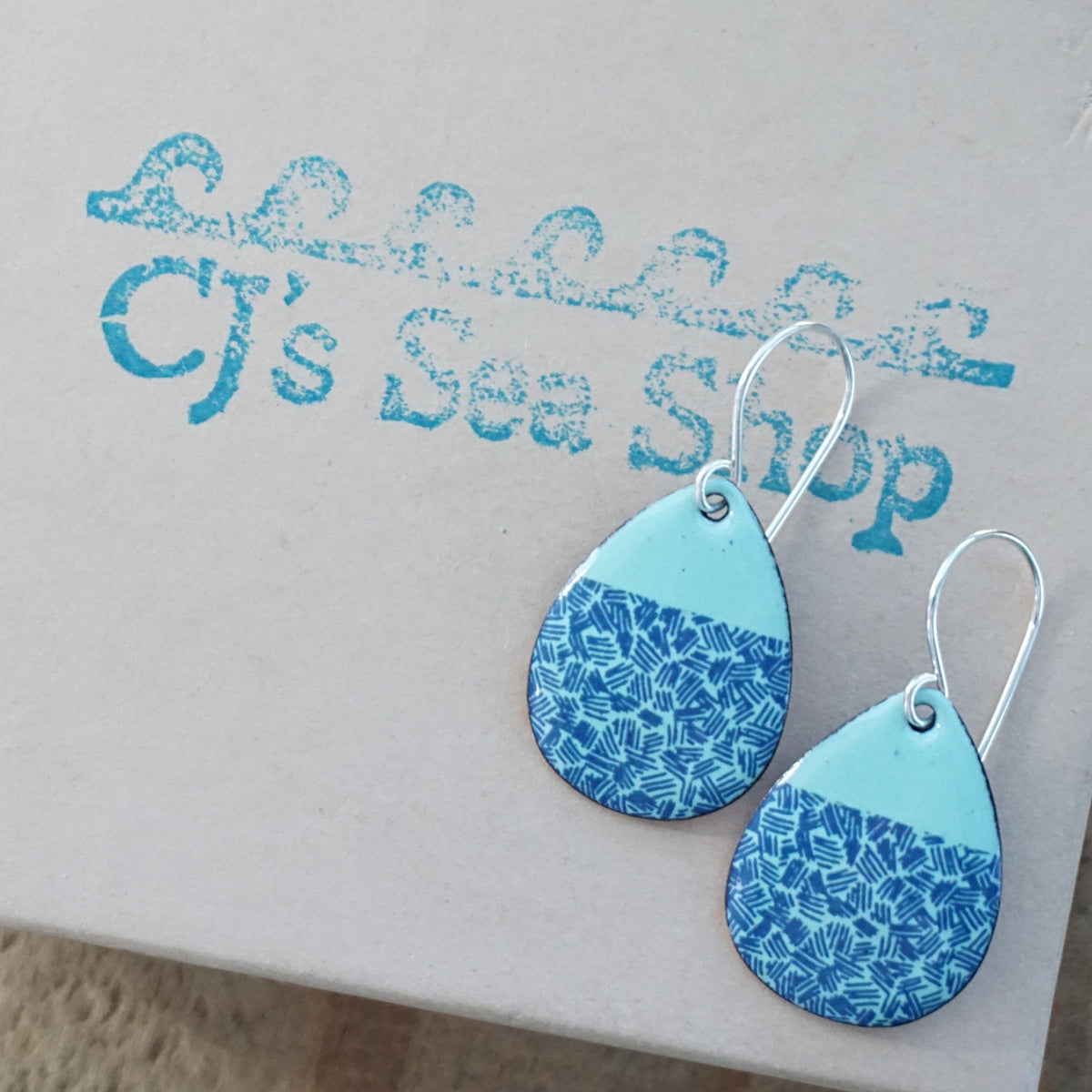 a pair of seafoam green enamel teardrop shaped earrings with blue accents, displayed on a gift box