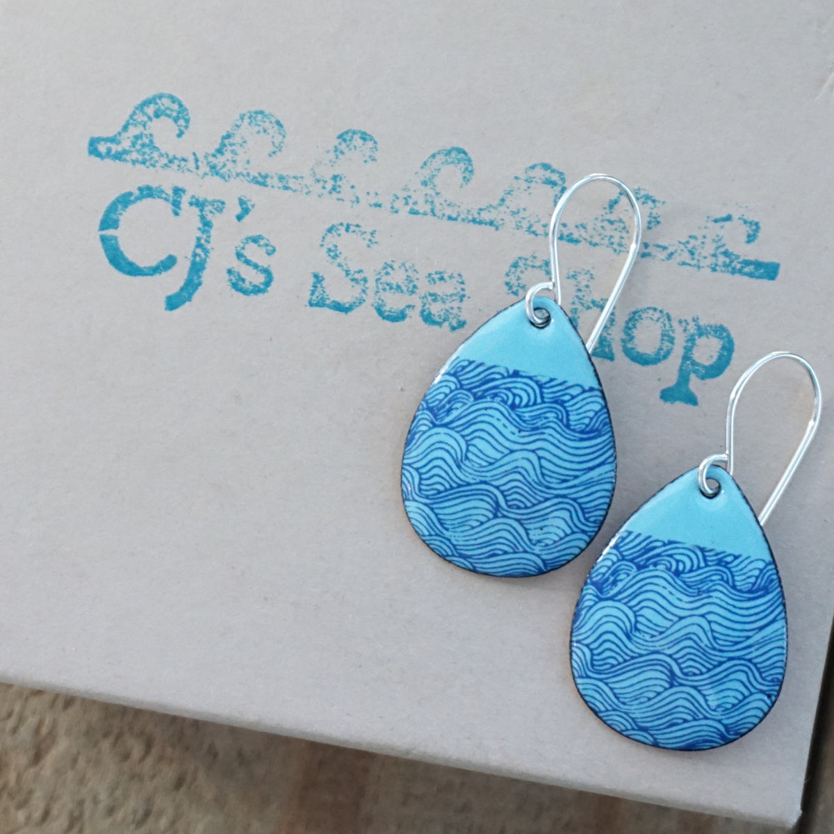 a pair of light blue enamel teardrop shaped earrings with blue wave accents, displayed on a gift box