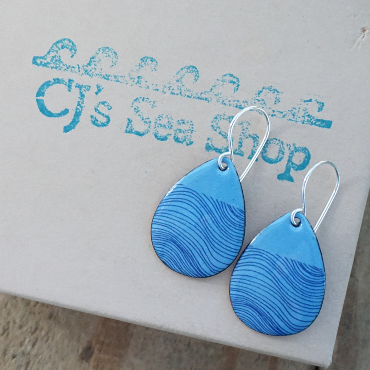 a pair of blue enamel teardrop shaped earrings with blue wavy line accents, displayed on a gift box