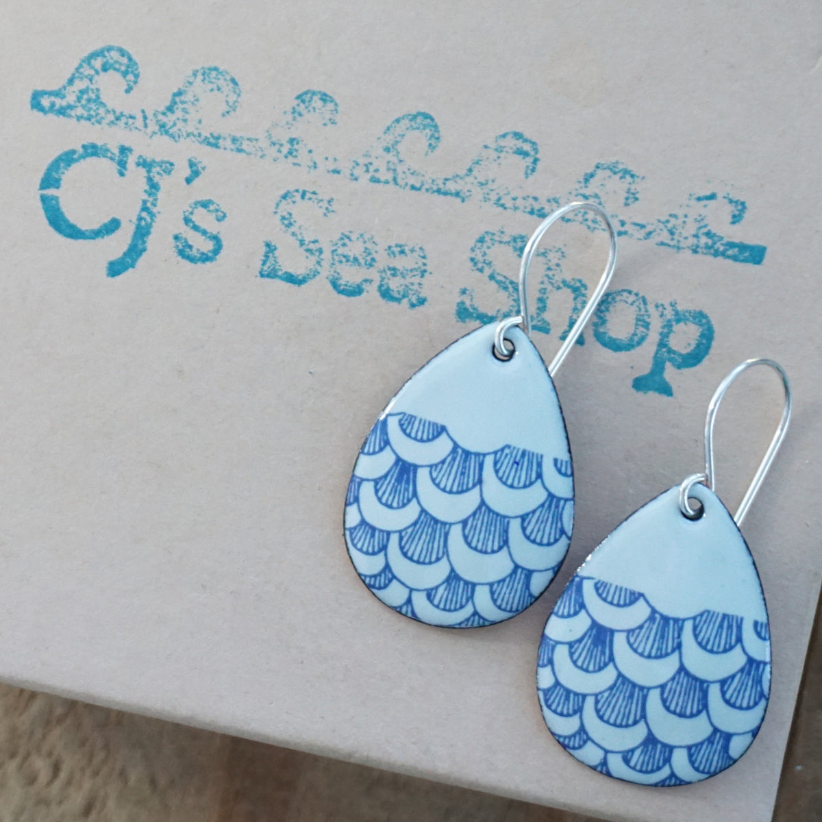 a pair of seafoam green enamel teardrop shaped earrings with blue fish scale accents, displayed on a gift box