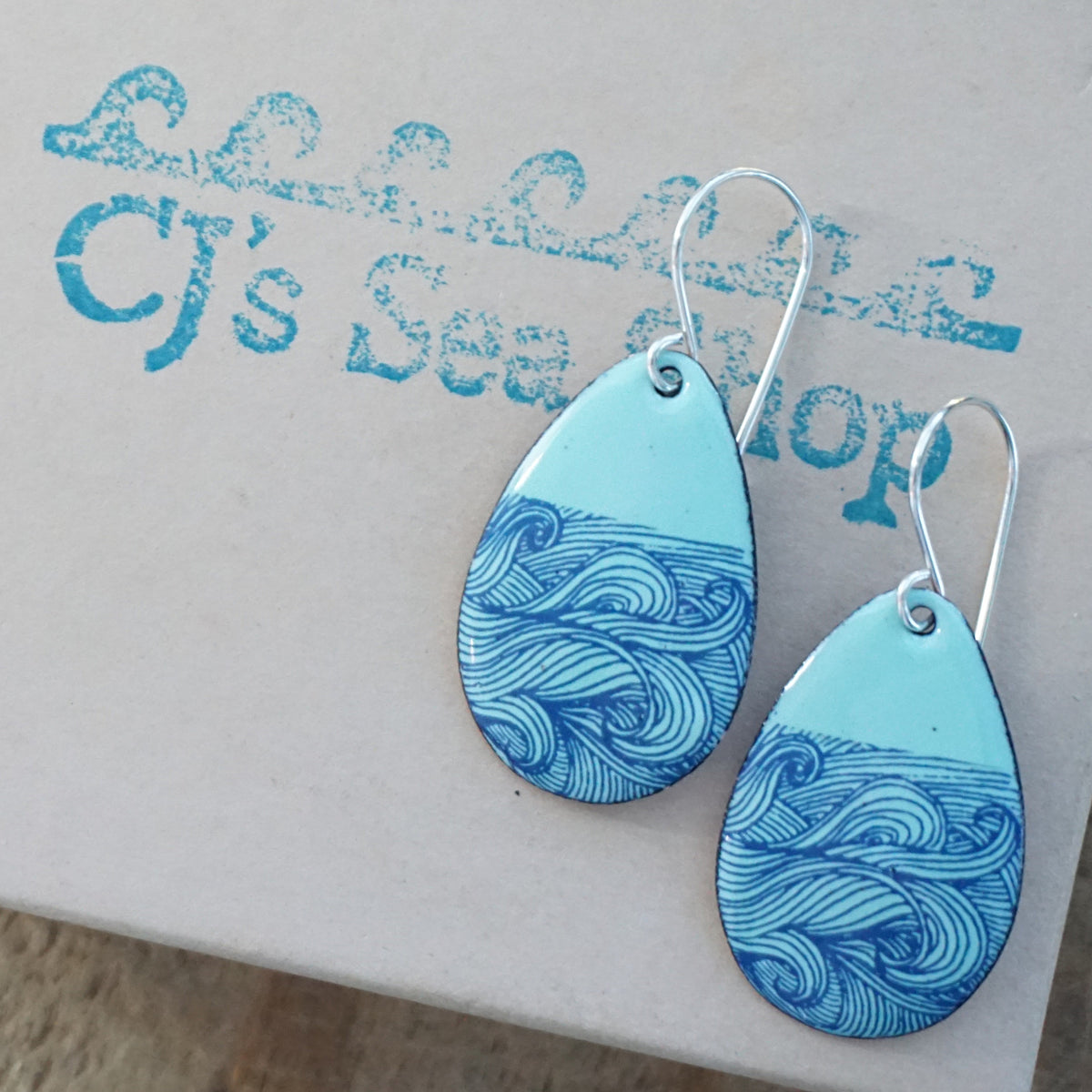 a pair of seafoam green enamel teardrop shaped earrings with blue wave accents, displayed on a gift box