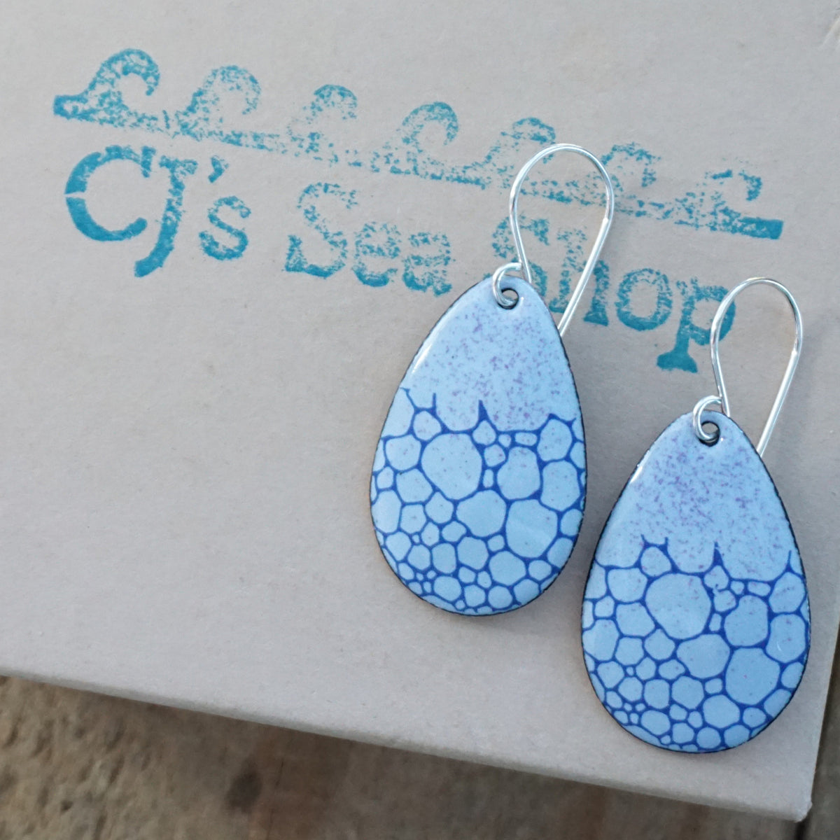 a pair of light purple enamel teardrop shaped earrings with blue bubble accents, displayed on a gift box