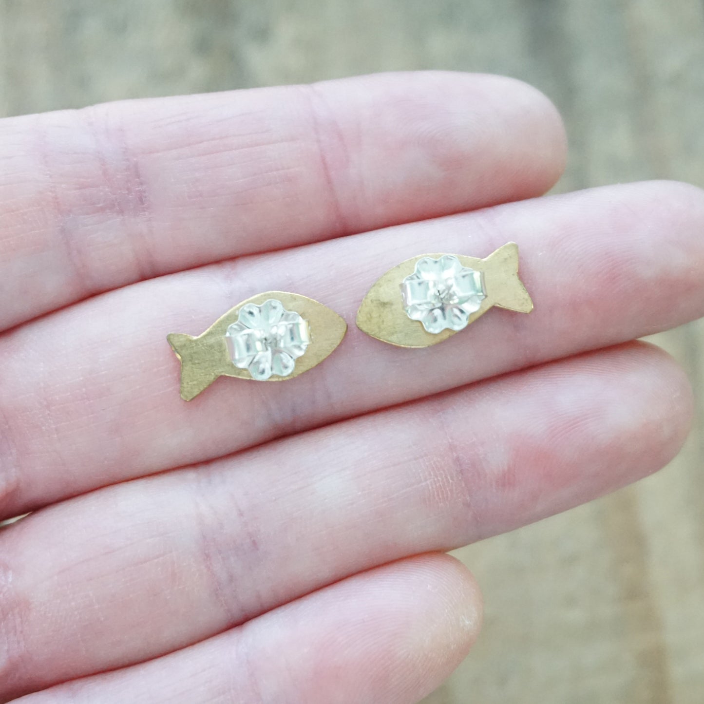 back view of a pair of Gold fish-shaped earrings held between fingers against a blurred background