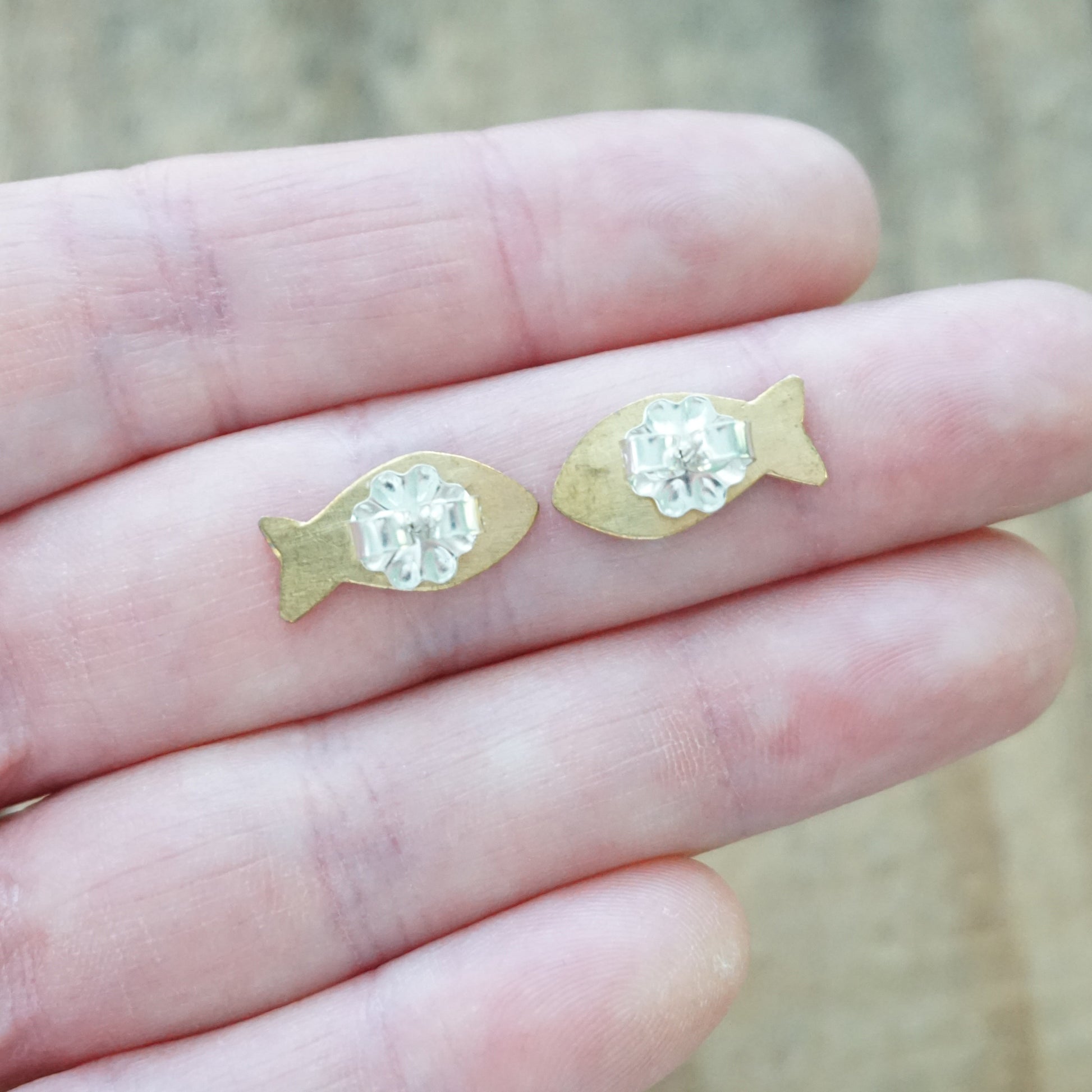 back view of a pair of Gold fish-shaped earrings held between fingers against a blurred background