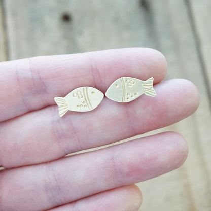Gold fish-shaped earrings held between fingers against a blurred wooden background