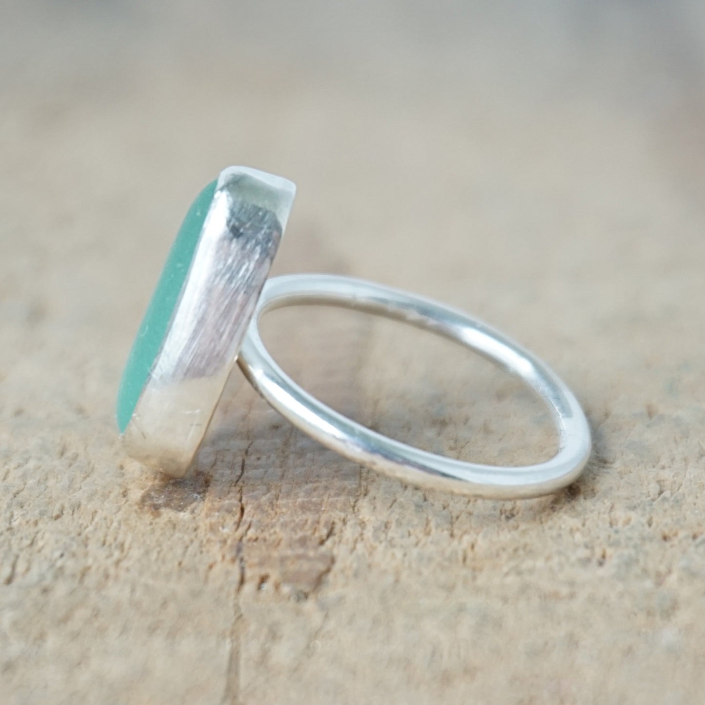 side view of a teal sea glass ring with a sterling silver setting, against a wooden background