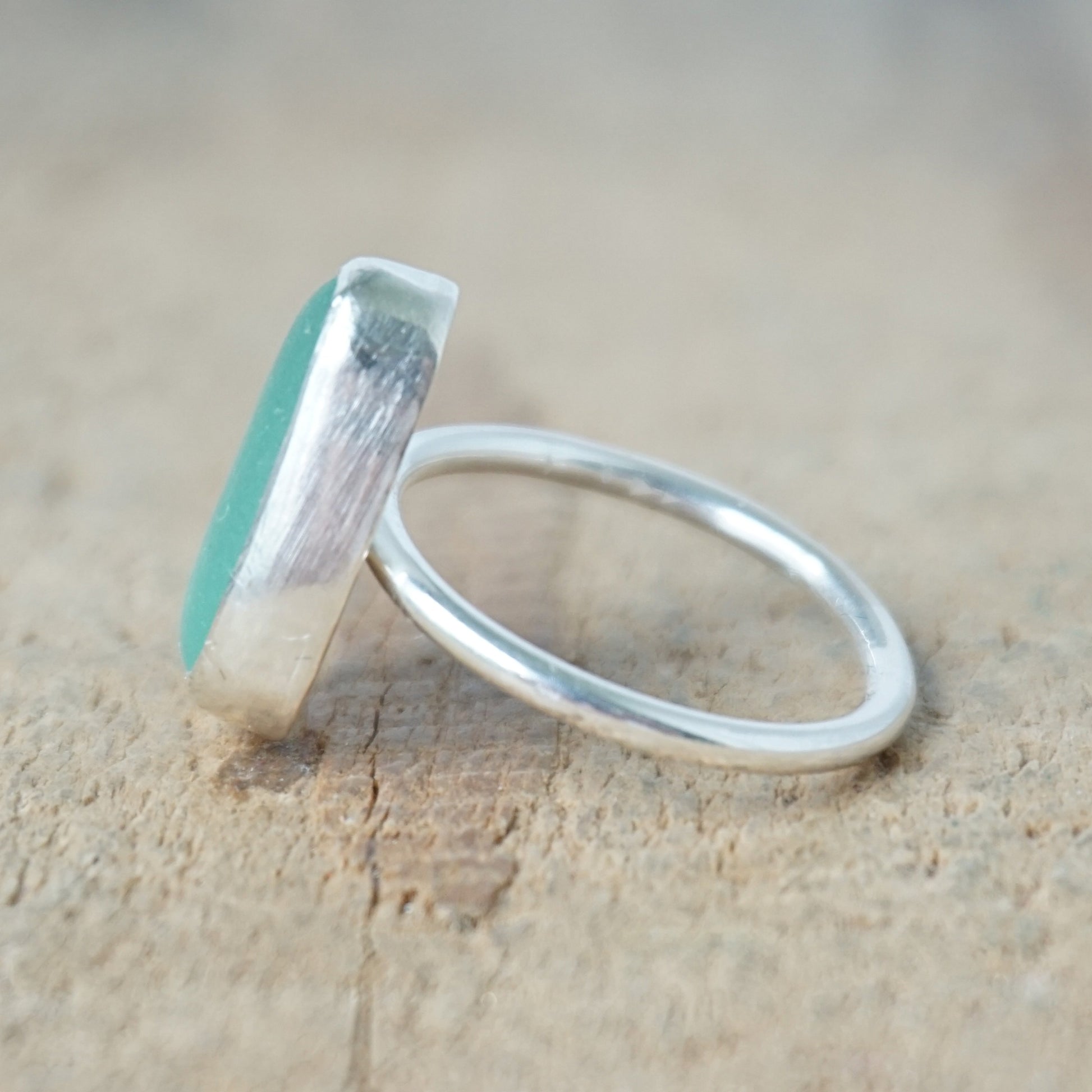 side view of a teal sea glass ring with a sterling silver setting, against a wooden background