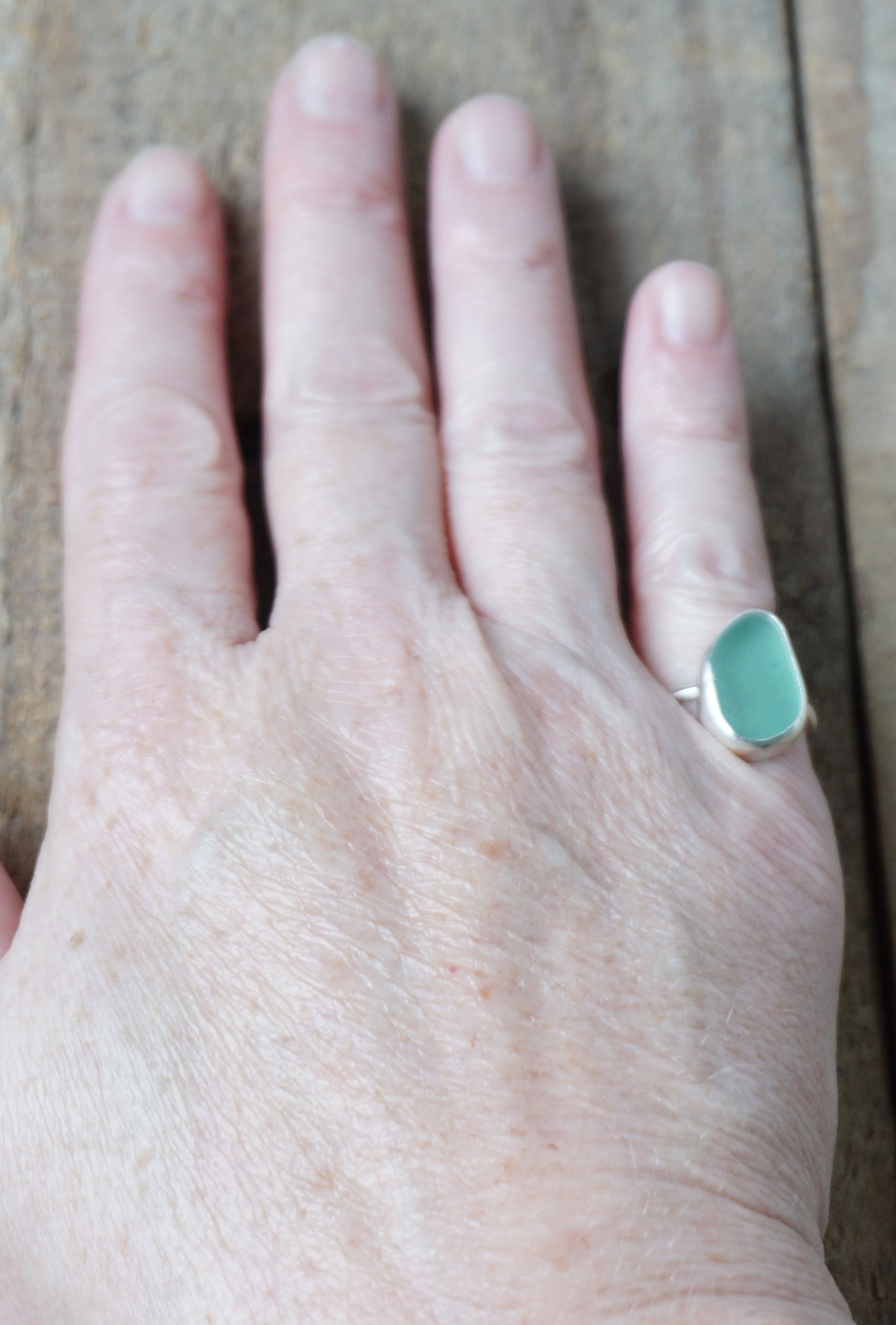 hand wearing a teal sea glass ring with a sterling silver setting, against a wooden background