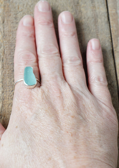 hand wearing a teal sea glass ring with a sterling silver setting, against a wooden background