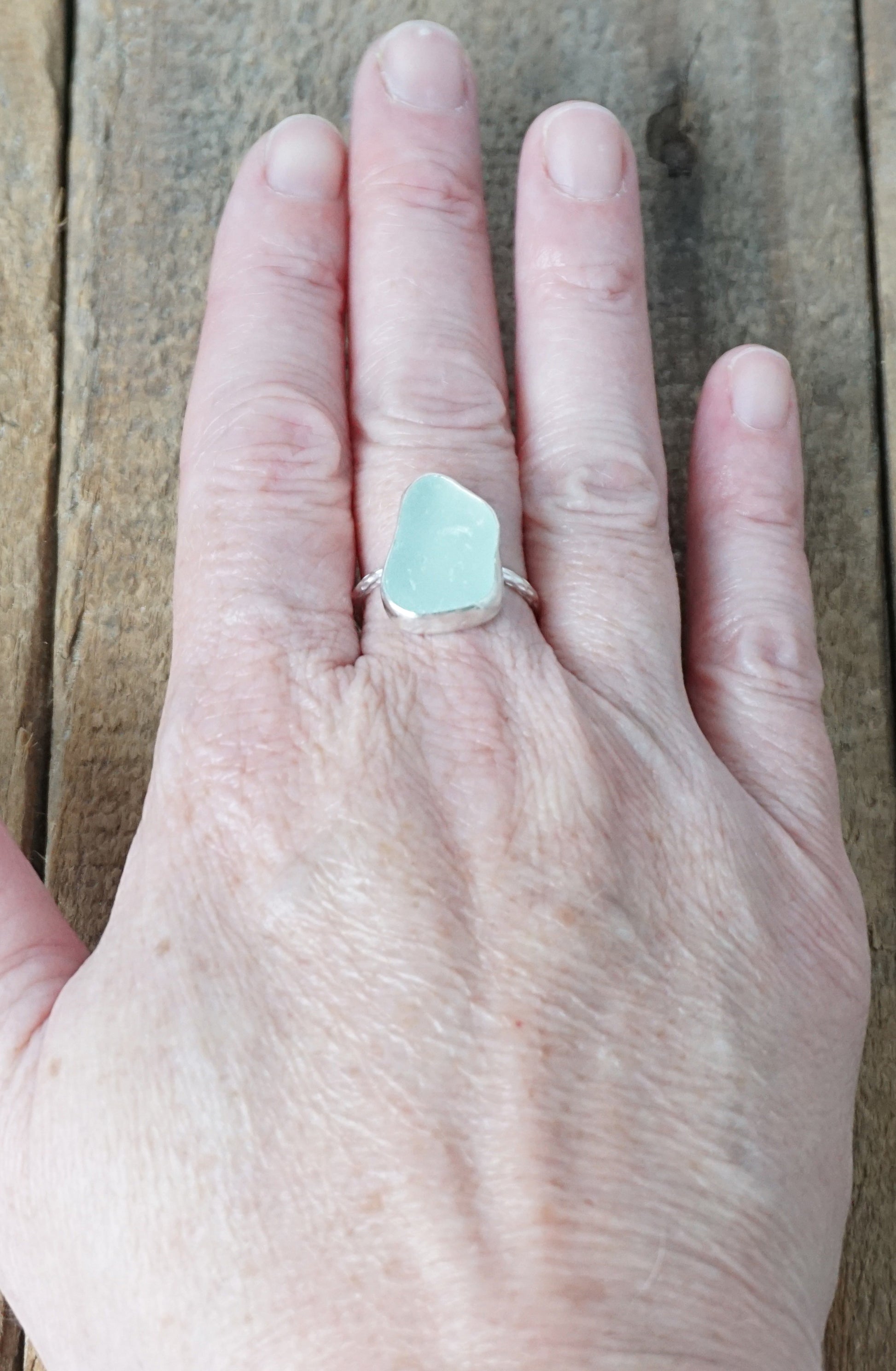 hand wearing a light aqua blue sea glass ring with a sterling silver setting, against a wooden background