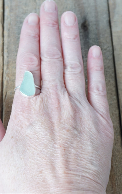 hand wearing a light aqua blue sea glass ring with a sterling silver setting, against a wooden background